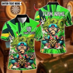 May include: Green polo shirt with a cartoon monkey smoking a cigar and holding a dartboard. The shirt features the number "420" and the text "TEAM NAME" and "YOUR NAME". The shirt has a black collar and is decorated with green and yellow patterns.