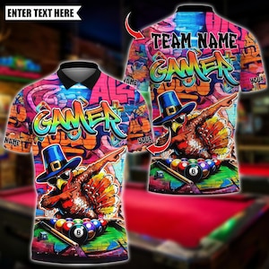 May include: A short-sleeved polo shirt featuring a vibrant graffiti-style background with the word GAMER in large colourful letters. A turkey wearing a blue pilgrim hat and sunglasses is dabbing over a pool table with billiard balls and a cue.