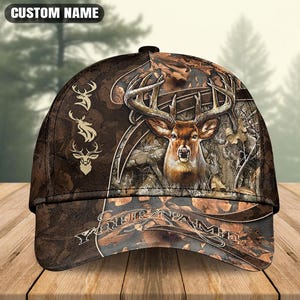 Könnte beinhalten: Eine braune Camouflage-Baseballkappe mit einer detaillierten Hirschkopf-Grafik, drei kleineren Hirschsilhouetten auf der linken Seite sowie dem Text CUSTOM NAME oben und YOUR NAME auf dem Schirm in weißer Schrift.