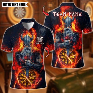 Puede incluir: Polo negro con un diseño de guerrero vikingo en llamas, sosteniendo una espada sobre una diana. La camisa incluye el texto "TEAM NAME" y espacio para "YOUR NAME". La parte superior tiene un gráfico de diana.