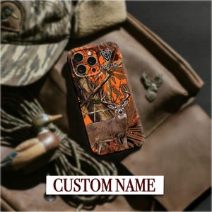 Könnte beinhalten: Handyhülle mit einem Hirschdesign und dem Text "YOUR NAME" auf einem Camouflage-Hintergrund. Die Hülle hat ein orange-braunes Farbschema, mit dem Text "CUSTOM NAME" unten.