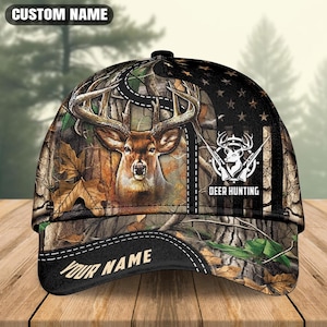 May include: A baseball cap featuring a camouflage pattern with a deer head, a stylised American flag design, and the text "DEER HUNTING" and "YOUR NAME". The cap has a structured crown and a curved brim with a black border and white stitching.