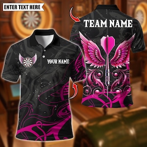 May include: Black polo shirt with a dartboard and wing design. The shirt features a pink and black color scheme with the text "YOUR NAME" and "TEAM NAME". The design includes a dart with pink wings.