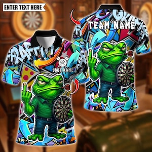 Op de afbeelding: Een poloshirt met korte mouwen met een cartoonkikker die een dartbord en een dart in zijn mond vasthoudt. De shirt heeft een kleurrijk graffiti-achtig ontwerp met de woorden "GRAFFITI" en "TEAM NAME". De kikker steekt zijn middelvinger op.