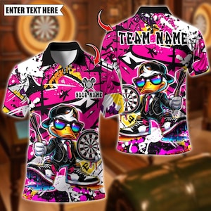 Op de afbeelding: Een poloshirt met een felroze en wit graffiti-patroon met een cartoon-eend in een pak, stropdas, zonnebril en pet. Het ontwerp bevat een dartbord, pijlen en tekstplaatsaanduidingen met ENTER TEXT HERE, TEAM NAME en YOUR NAME.