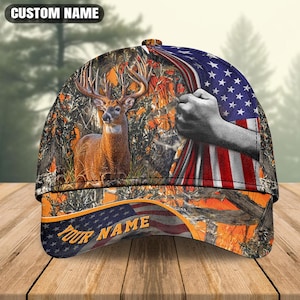 Può includere: Un cappellino da baseball con un cervo in un ambiente boschivo, una mano che afferra una bandiera americana e motivi mimetici. Il testo CUSTOM NAME appare in alto e il testo YOUR NAME è stampato in giallo sulla visiera sopra un disegno di bandiera.