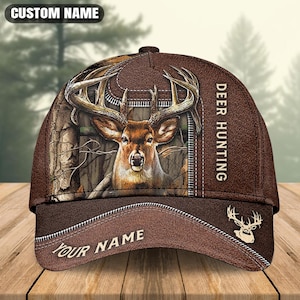 May include: A brown baseball cap featuring a deer head graphic with large antlers, a camouflage forest pattern, and a zipper design. The text DEER HUNTING is printed vertically on the side, while CUSTOM NAME and YOUR NAME appear as placeholders.