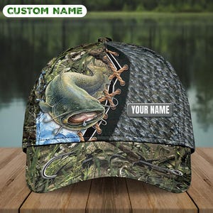 Op de afbeelding: Een baseballpet met een afbeelding van een meerval in het water, een vishaak, een grijs paneel met textuur en een camouflagepatroon. De pet bevat een rechthoekige patch met de tekst YOUR NAME en een CUSTOM NAME label aan de bovenzijde.