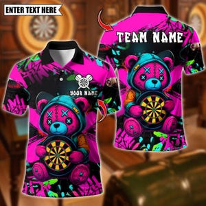 May include: Pink polo shirt featuring a cartoon bear holding a dartboard. The bear has a blue hood and stitched details. The shirt has the text "TEAM NAME" and "YOUR NAME" with a dartboard graphic. The words "ENTER TEXT HERE" are also visible.