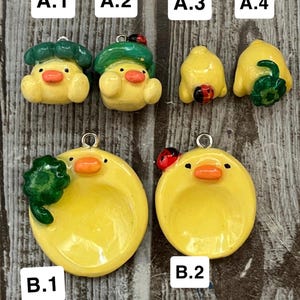 May include: A collection of yellow duck-shaped charms with orange beaks and black eyes. Some charms have green hats or accessories, and a few have red and black ladybug accents. The charms are designed for jewelry making or crafting.