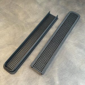 Può includere: Due griglie di ventilazione in plastica nera con un motivo a griglia. Le griglie di ventilazione sono rettangolari e hanno un bordo leggermente curvo.