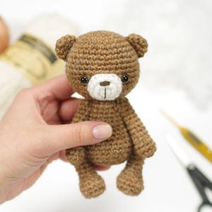 Op de afbeelding: Een kleine, handgemaakte bruine gehaakte teddybeer met een witte snuit en zwarte ogen. De beer wordt in een hand gehouden, wat de gedetailleerde steken en het compacte formaat laat zien. Een charmant, handgemaakt speelgoed.