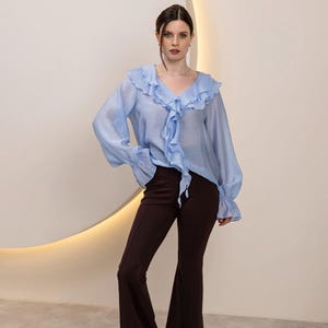 May include: A light blue blouse with ruffled trim and long sleeves, paired with dark brown flared pants. The blouse features a V-neck and a tie detail. The pants have a high waist and a wide leg. The outfit is set against a neutral background.