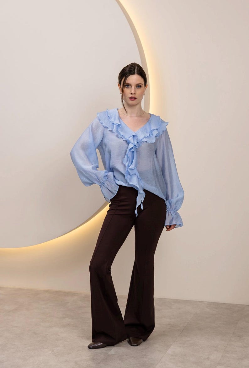 May include: A light blue blouse with ruffled trim and long sleeves, paired with dark brown flared pants. The blouse features a V-neck and a tie detail. The pants have a high waist and a wide leg. The outfit is set against a neutral background.