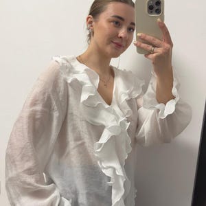 May include: A white blouse with a ruffled neckline and long sleeves. The blouse has a V-neck and a flowing, lightweight fabric. The sleeves have a ruffled detail at the cuffs. The blouse is paired with black pinstripe pants.