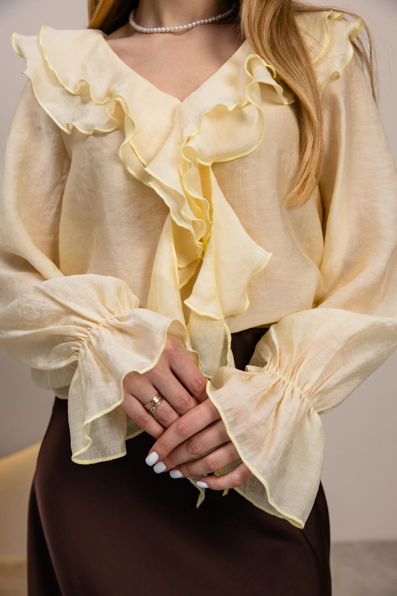 May include: A cream-colored blouse with a V-neck and ruffled trim in a contrasting yellow. The long sleeves have gathered cuffs with matching yellow trim. The blouse is paired with a brown skirt. Fashion apparel.