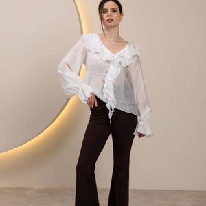May include: A woman wearing a white blouse with ruffled details and long sleeves, paired with dark brown flared pants. The blouse has a V-neck and the pants are high-waisted. The background is a neutral color with a circular design.