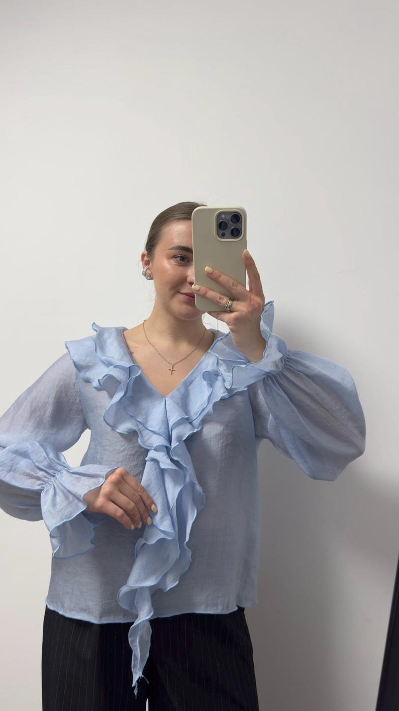 May include: Light blue blouse with ruffled detailing around the neckline and sleeves. The blouse has long sleeves with a gathered cuff. The fabric appears to be lightweight and semi-sheer. The blouse is paired with black pinstripe pants.