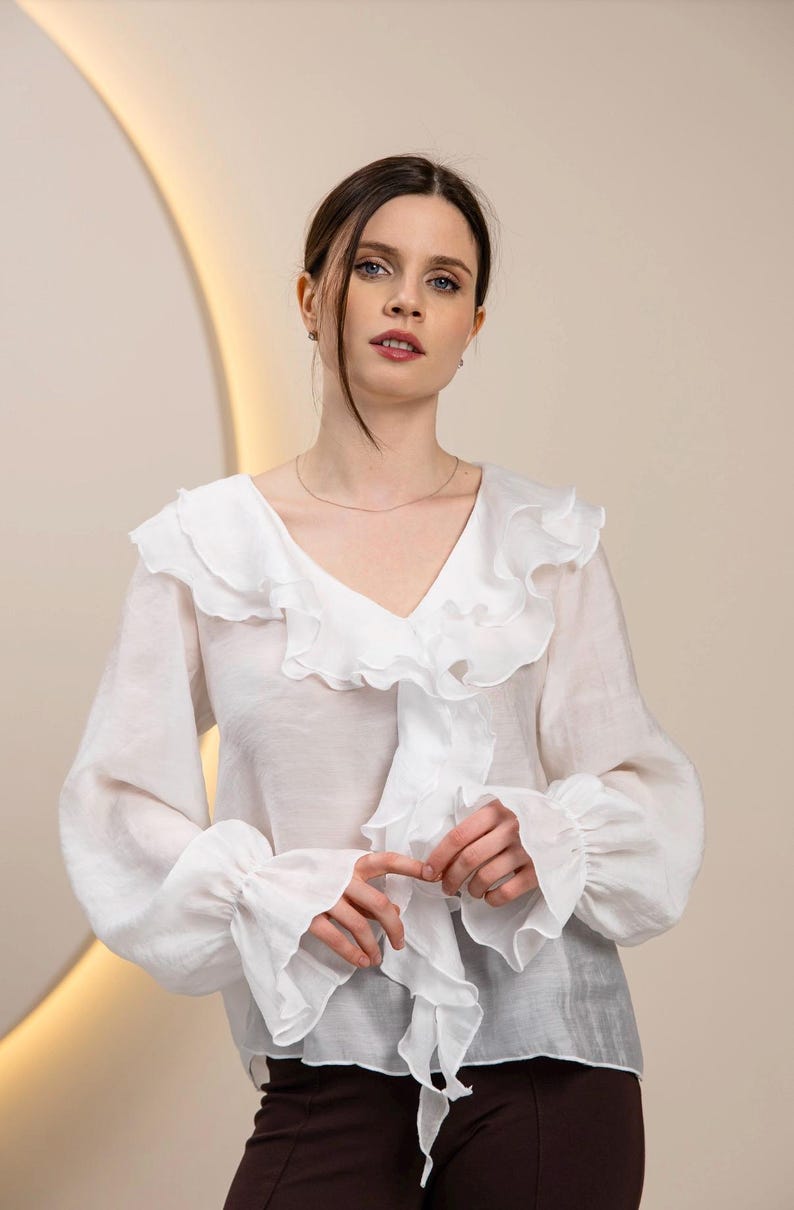 May include: White blouse with a V-neck and ruffled collar. The long-sleeved top features ruffled cuffs and a flowing, lightweight fabric. The blouse is a classic style, suitable for various occasions.