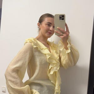 May include: A pale yellow blouse with a ruffled neckline and sleeves. The blouse has long sleeves with ruffled cuffs. The fabric appears to be lightweight and slightly sheer. The blouse is paired with black pinstripe pants.