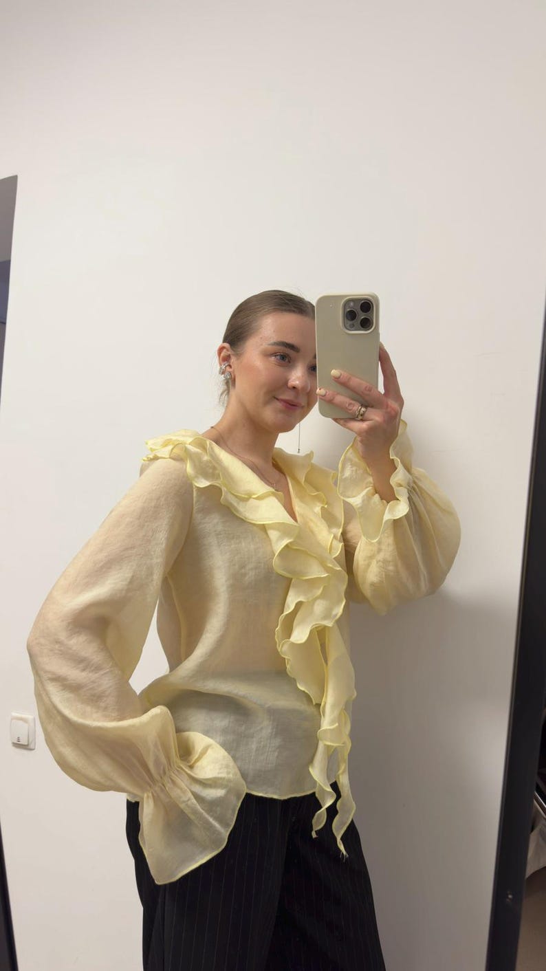 May include: A pale yellow blouse with a ruffled neckline and sleeves. The blouse has long sleeves with ruffled cuffs. The fabric appears to be lightweight and slightly sheer. The blouse is paired with black pinstripe pants.