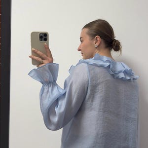 May include: A light blue blouse with ruffled collar and cuffs. The long-sleeved top has a sheer, flowing fabric. The blouse is paired with black and white striped pants. The person is holding a phone.