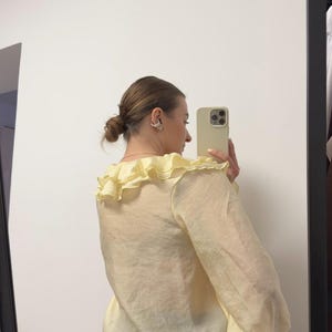 May include: Pale yellow blouse with a ruffled collar and long, billowy sleeves. The sheer fabric creates a light and airy look. The blouse is paired with black pinstripe pants. The image is taken in front of a mirror.