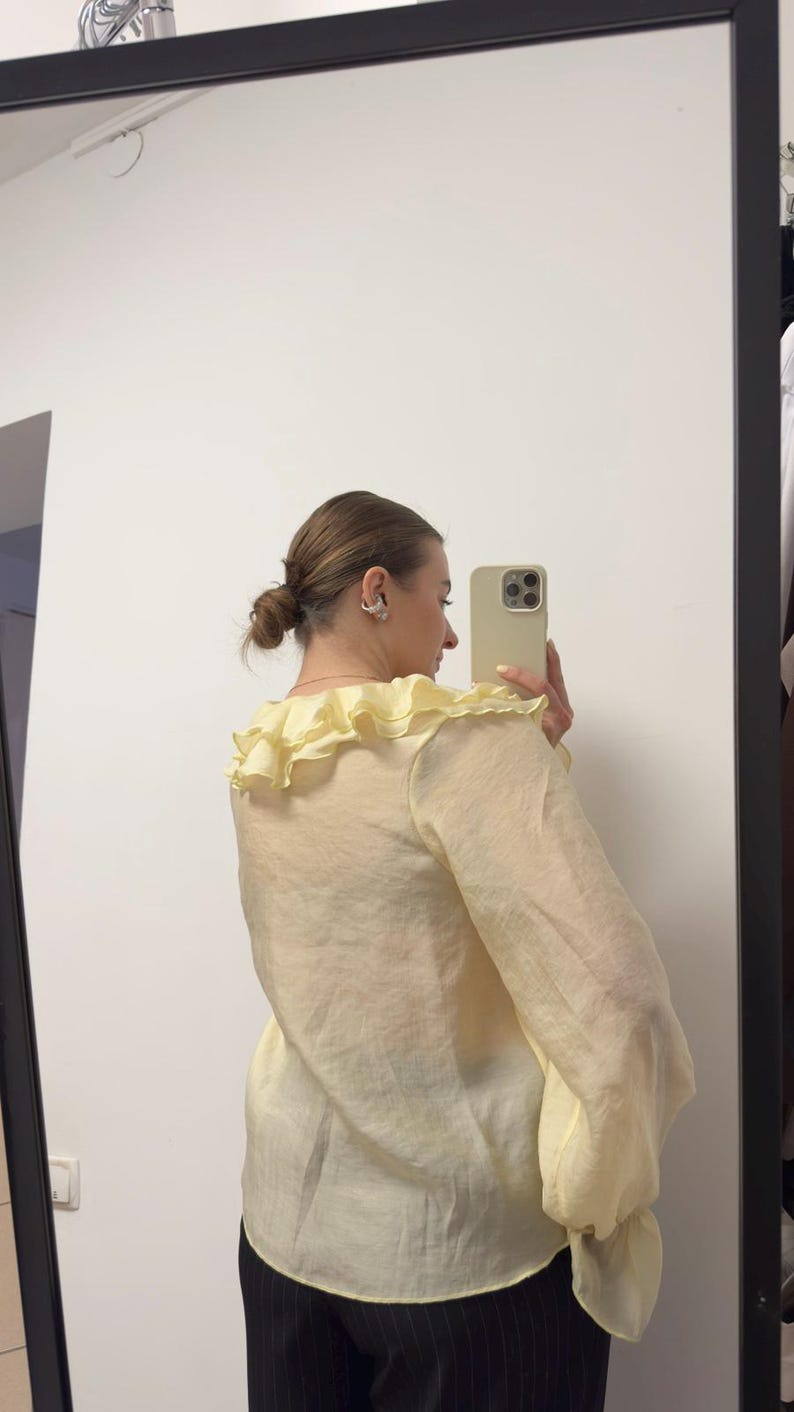 May include: Pale yellow blouse with a ruffled collar and long, billowy sleeves. The sheer fabric creates a light and airy look. The blouse is paired with black pinstripe pants. The image is taken in front of a mirror.
