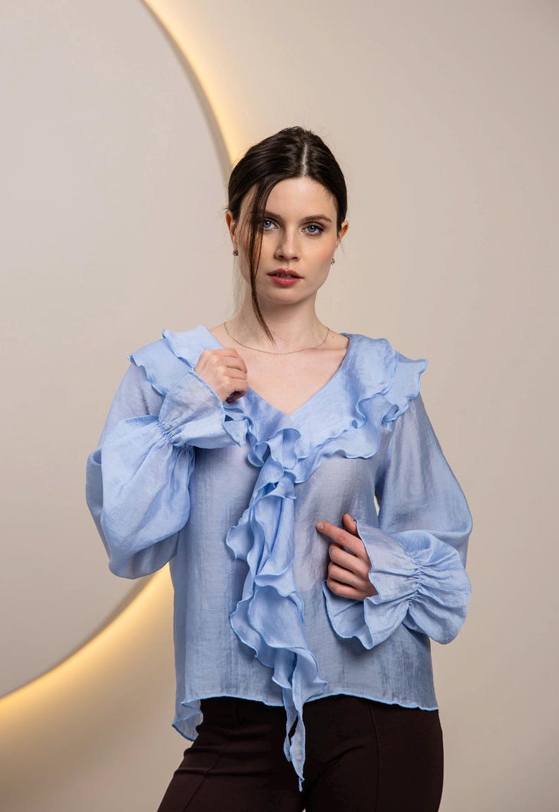 May include: A light blue blouse with a V-neck and ruffled trim. The long sleeves have ruffled cuffs. The blouse is made of a lightweight fabric.