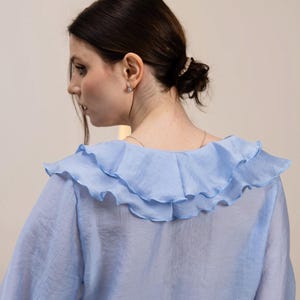 May include: A light blue blouse with a ruffled collar. The blouse has long sleeves and a sheer, flowing fabric. The ruffled collar adds a decorative element to the top. The blouse is a solid color and appears to be made of a lightweight material.