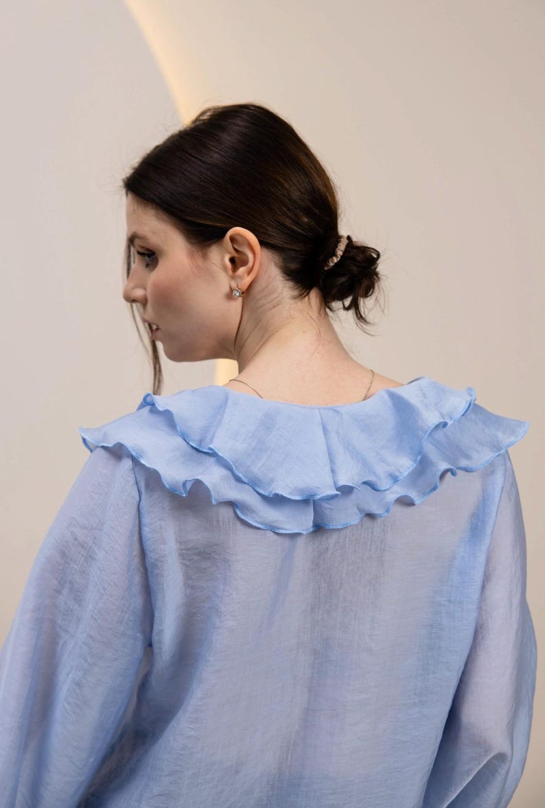 May include: A light blue blouse with a ruffled collar. The blouse has long sleeves and a sheer, flowing fabric. The ruffled collar adds a decorative element to the top. The blouse is a solid color and appears to be made of a lightweight material.