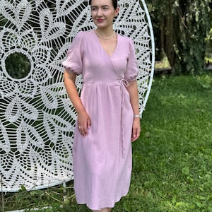 May include: A light pink midi-length wrap dress featuring a V-neckline, short puff sleeves with gathered cuffs, and a side tie closure. The garment is made of a textured fabric and falls to a mid-calf hemline, providing a simple and clean silhouette.