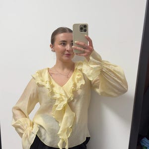 May include: A pale yellow blouse with a ruffled front and long, billowy sleeves. The blouse has a V-neck and is paired with black pinstripe pants. The image is taken in front of a mirror.