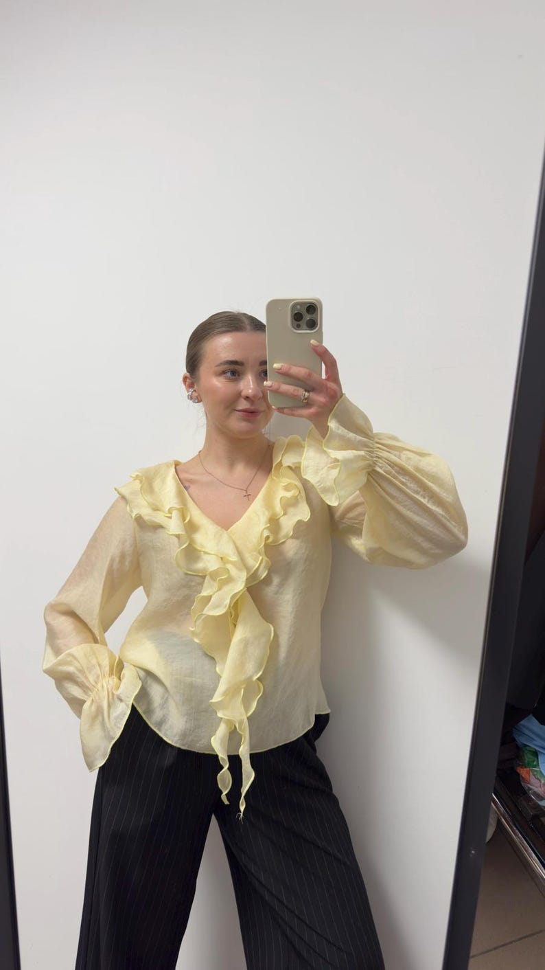 May include: A pale yellow blouse with a ruffled front and long, billowy sleeves. The blouse has a V-neck and is paired with black pinstripe pants. The image is taken in front of a mirror.