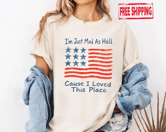 May include: Cream-colored t-shirt featuring a stylized American flag design with red stripes and blue stars. The text "I'm Just Mad As Hell Cause I Loved This Place" is printed below the flag. The shirt is part of a casual wear collection.