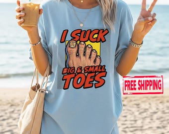 May include: Light blue t-shirt with the text "I SUCK BIG & SMALL TOES" in red and white. The graphic features a cartoon foot with a yellow background and black halftone dots. The shirt is a casual style.
