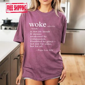 May include: A mauve-colored t-shirt with the word "woke" printed in white, along with a definition and quote. The text includes the pronunciation, a definition, and a quote by Pope Leo XIV. The shirt has short sleeves.