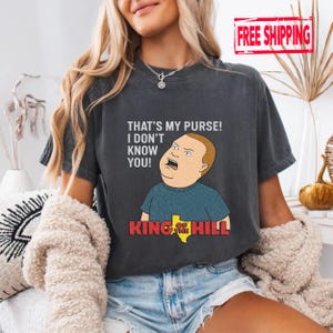 May include: Dark grey t-shirt featuring a cartoon image of Bobby Hill from King of the Hill, with the quote "THAT'S MY PURSE! I DON'T KNOW YOU!". The shirt has a distressed look.