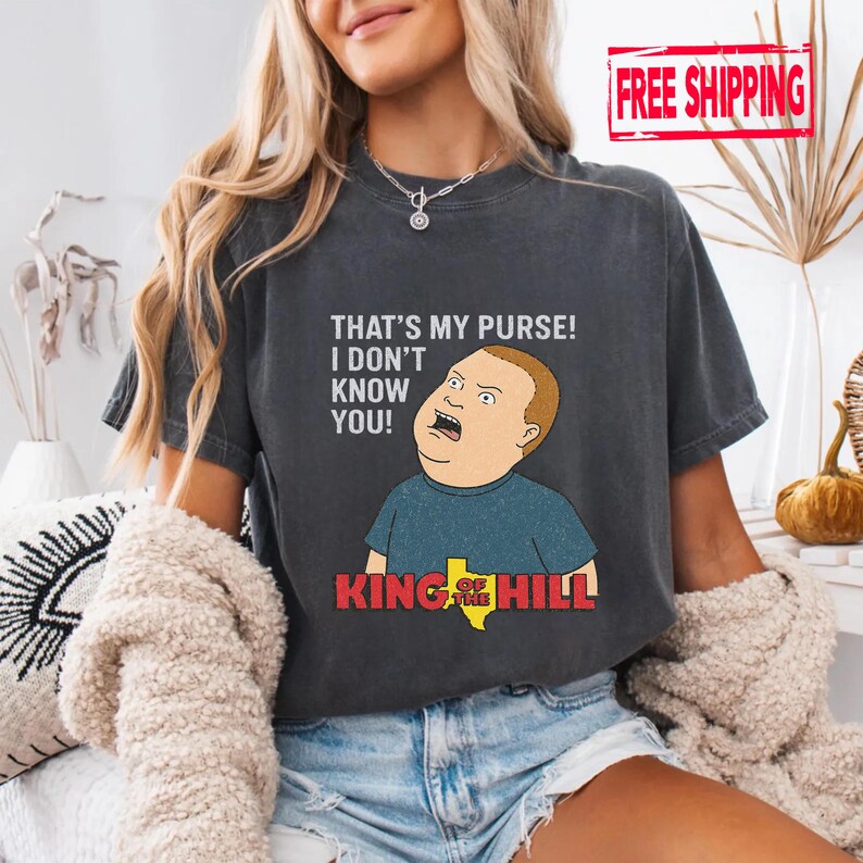 May include: Dark grey t-shirt featuring a cartoon image of Bobby Hill from King of the Hill, with the quote "THAT'S MY PURSE! I DON'T KNOW YOU!". The shirt has a distressed look.