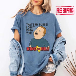 May include: Light blue t-shirt with a cartoon image of Bobby Hill from King of the Hill, displaying the quote "THAT'S MY PURSE! I DON'T KNOW YOU!" and the show's logo. The shirt is a light blue colour.