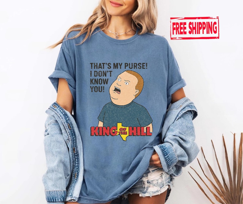 May include: Light blue t-shirt with a cartoon image of Bobby Hill from King of the Hill, displaying the quote "THAT'S MY PURSE! I DON'T KNOW YOU!" and the show's logo. The shirt is a light blue colour.