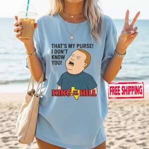May include: Pale blue t-shirt with a cartoon image of Bobby Hill from King of the Hill, and the quote "THAT'S MY PURSE! I DON'T KNOW YOU!" and the show's logo. The shirt has a loose fit.