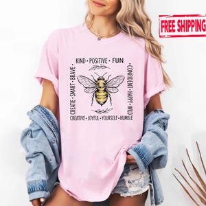 May include: A light pink t-shirt featuring a central graphic of a yellow and black bee surrounded by a square border of text including the words KIND, POSITIVE, FUN, CONFIDENT, HAPPY, WILD, HUMBLE, YOURSELF, JOYFUL, CREATIVE, SMART, and BRAVE.