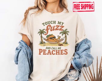 May include: A cream-coloured short-sleeve t-shirt featuring a graphic of an orange peach wearing sunglasses, relaxing in a hammock between two palm trees, holding a drink. The text reads TOUCH MY fuzz AND CALL ME PEACHES.