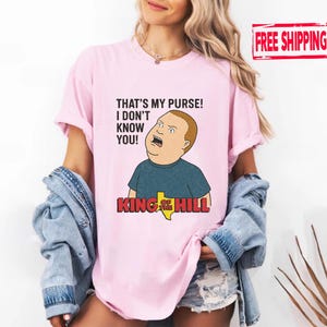 May include: Pale pink t-shirt with a cartoon image of Bobby Hill from King of the Hill. The shirt displays the quote "THAT'S MY PURSE! I DON'T KNOW YOU!" and the show's logo. The t-shirt is worn by a person.
