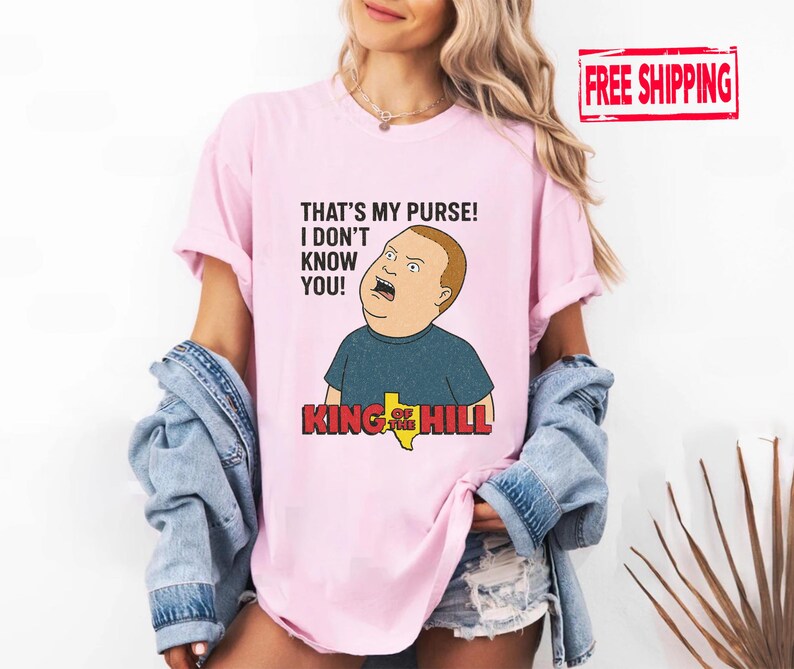 May include: Pale pink t-shirt with a cartoon image of Bobby Hill from King of the Hill. The shirt displays the quote "THAT'S MY PURSE! I DON'T KNOW YOU!" and the show's logo. The t-shirt is worn by a person.