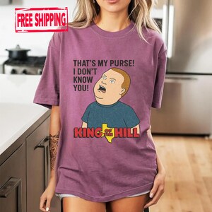 May include: A mauve t-shirt with a cartoon image of Bobby Hill from King of the Hill. The graphic includes the phrase "THAT'S MY PURSE! I DON'T KNOW YOU!" and the show's logo. The shirt has short sleeves.