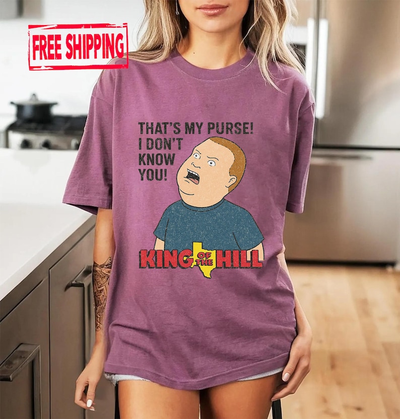 May include: A mauve t-shirt with a cartoon image of Bobby Hill from King of the Hill. The graphic includes the phrase "THAT'S MY PURSE! I DON'T KNOW YOU!" and the show's logo. The shirt has short sleeves.
