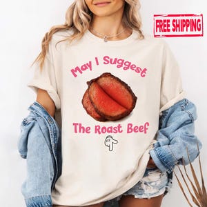 May include: Cream-colored t-shirt with the text "May I Suggest The Roast Beef" in pink. The graphic features a sliced roast beef image and a hand pointing down. The shirt is a casual, comfortable style.
