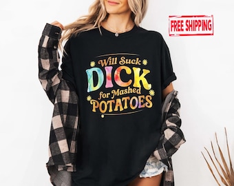 May include: Black t-shirt with the text "Will Suck Dick for Mashed Potatoes" in a colorful, tie-dye style font. The shirt is a crew neck and is being worn with a plaid shirt.